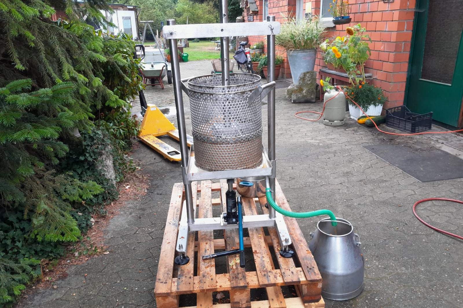 Die selbstgebaute Spindelpresse steht auf einer Palette im Hof, der Presskorb im Rahmen, daneben eine Milchkanne am grünen Schlauch; dahinter die Klinkerwand mit Sonnenblumen.