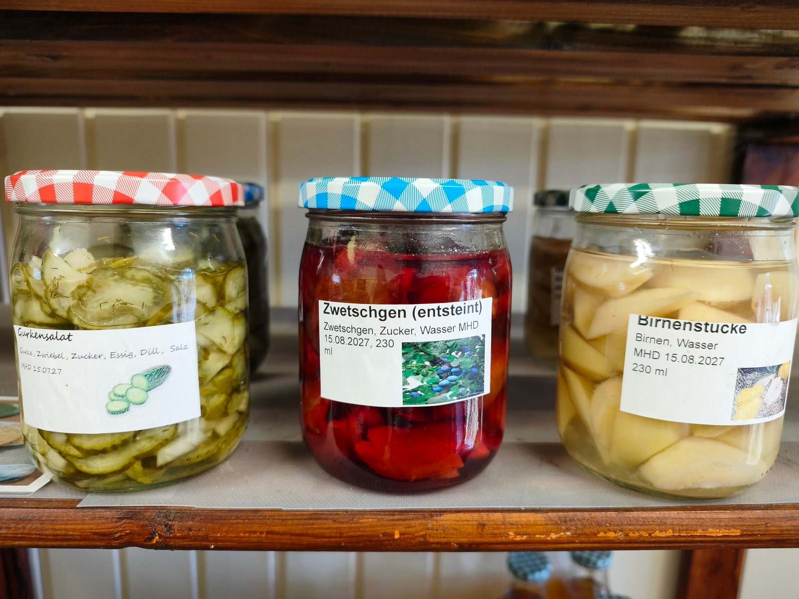 Drei große Gläser stehen nebeneinander auf einem Brett im Verkaufswagen — Gurkensalat, entsteinte Zwetschgen in dunkelrotem Saft und helle Birnenstücke, jedes mit kariertem Deckel und beschriftetem Etikett.