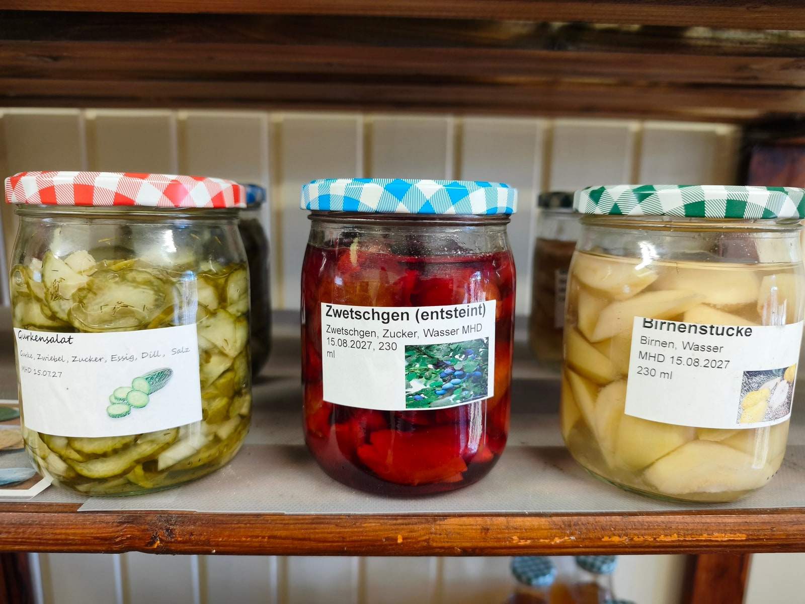 Drei große Gläser stehen nebeneinander auf einem Brett im Verkaufswagen — Gurkensalat, entsteinte Zwetschgen in dunkelrotem Saft und helle Birnenstücke, jedes mit kariertem Deckel und beschriftetem Etikett.