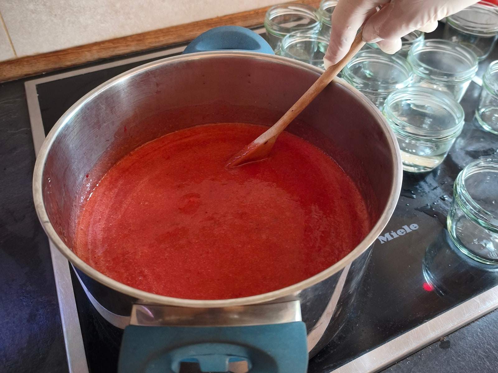 Ein großer Topf mit roter Marmelade steht auf dem Kochfeld, eine Hand im Handschuh rührt mit einer Holzkelle darin; rechts daneben warten leere Einmachgläser.