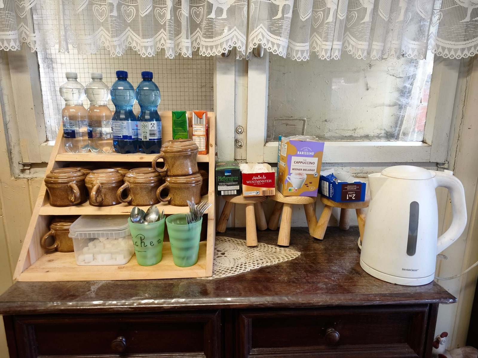 Die Getränkeecke im Verkaufswagen — auf einem Holzregal stehen tönerne Becher, Gläser mit Besteck und Wasserflaschen, daneben auf kleinen Hockern Kaffee- und Teepäckchen, rechts ein weißer Wasserkocher auf der alten Anrichte.