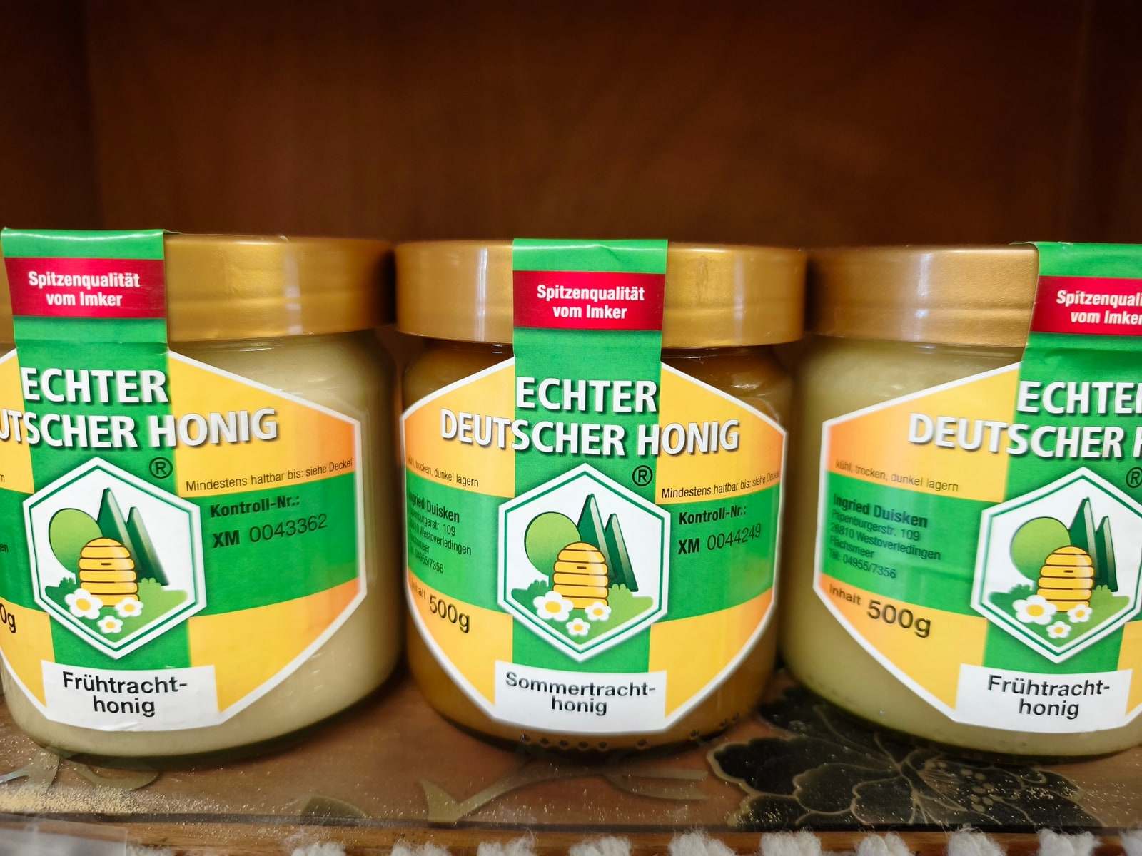 Drei Gläser Honig mit goldenen Deckeln stehen nebeneinander auf einem Brett, zwei davon mit hellem, cremig gerührtem Frühtrachthonig, das mittlere mit dunklerem Sommertrachthonig.