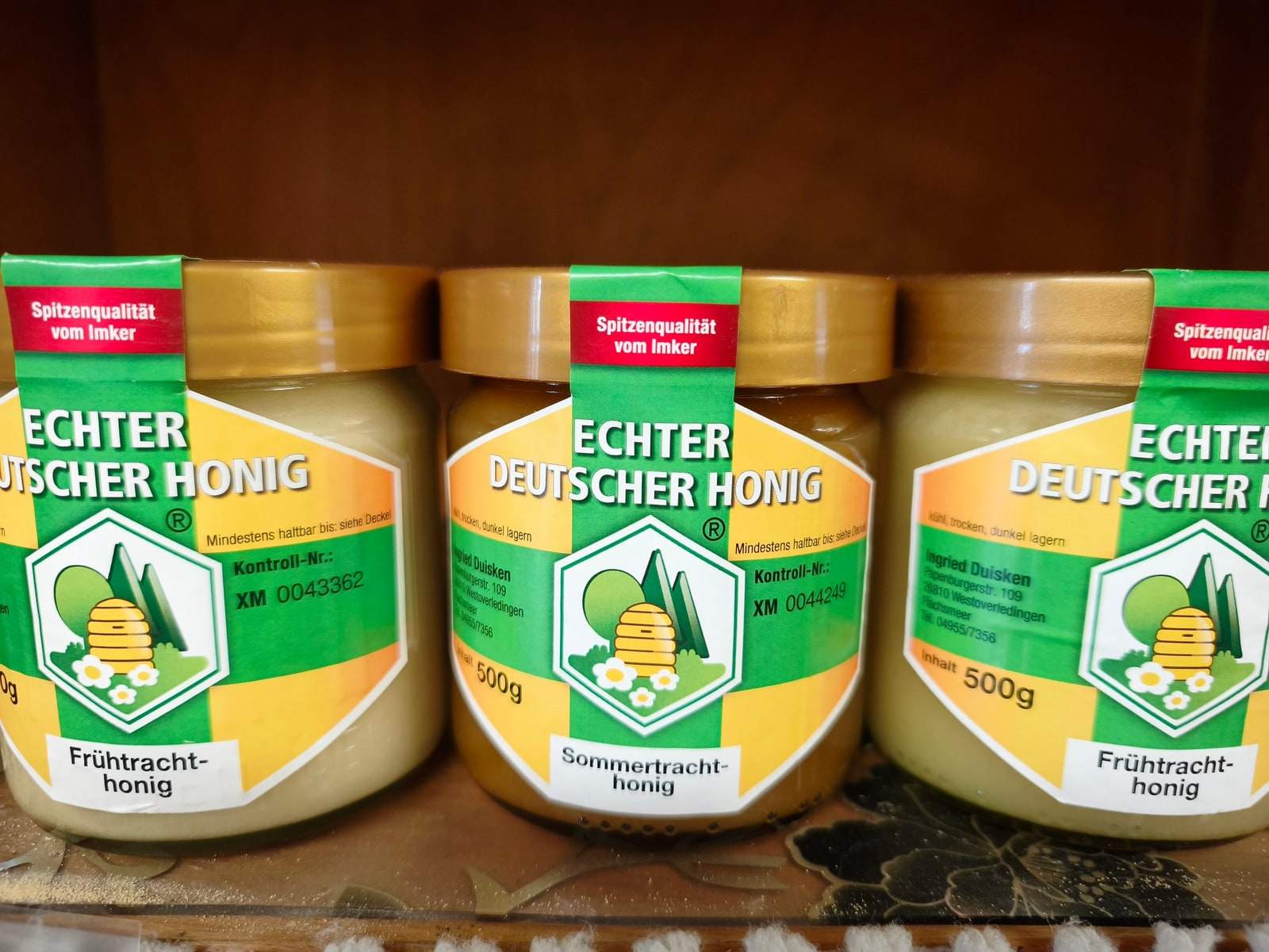 Drei Gläser Honig mit goldenen Deckeln stehen nebeneinander auf einem Brett, zwei davon mit hellem, cremig gerührtem Frühtrachthonig, das mittlere mit dunklerem Sommertrachthonig.