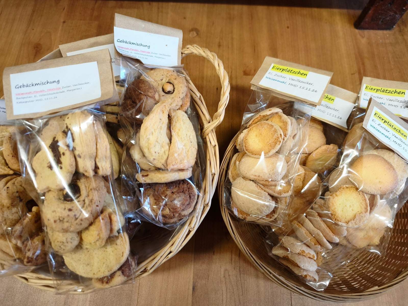 Zwei Weidenkörbe voller klarer Tüten mit Gebäck stehen auf einem Holzbrett, links dunklere Gebäckmischung, rechts helle Eierplätzchen; an jeder Tüte hängt ein beschriftetes Papierschild.
