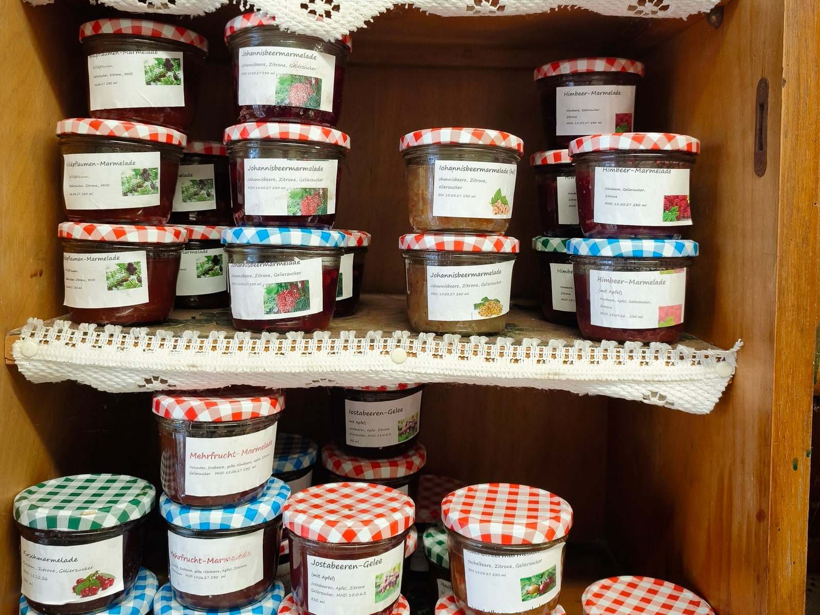 Zwei Bretter eines alten Küchenschranks, dicht gestapelt mit Gläsern Marmelade und Gelee in Rot, Blau und Gelb; die Deckel sind rot, blau und grün kariert, jedes Glas trägt ein beschriftetes Etikett mit Sorte und Zutaten.