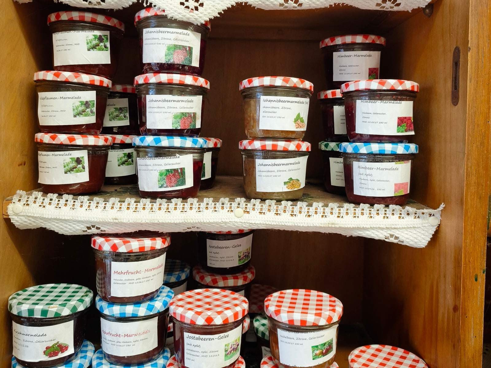 Zwei Bretter eines alten Küchenschranks, dicht gestapelt mit Gläsern Marmelade und Gelee in Rot, Blau und Gelb; die Deckel sind rot, blau und grün kariert, jedes Glas trägt ein beschriftetes Etikett mit Sorte und Zutaten.
