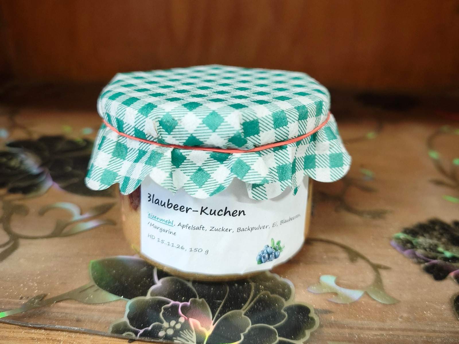 Ein Einmachglas mit Blaubeer-Kuchen steht auf einem gemusterten Brett im Verkaufswagen, über den Deckel ist ein grün-weiß kariertes Stoffdeckchen gebunden, davor ein handbeschriftetes Etikett.
