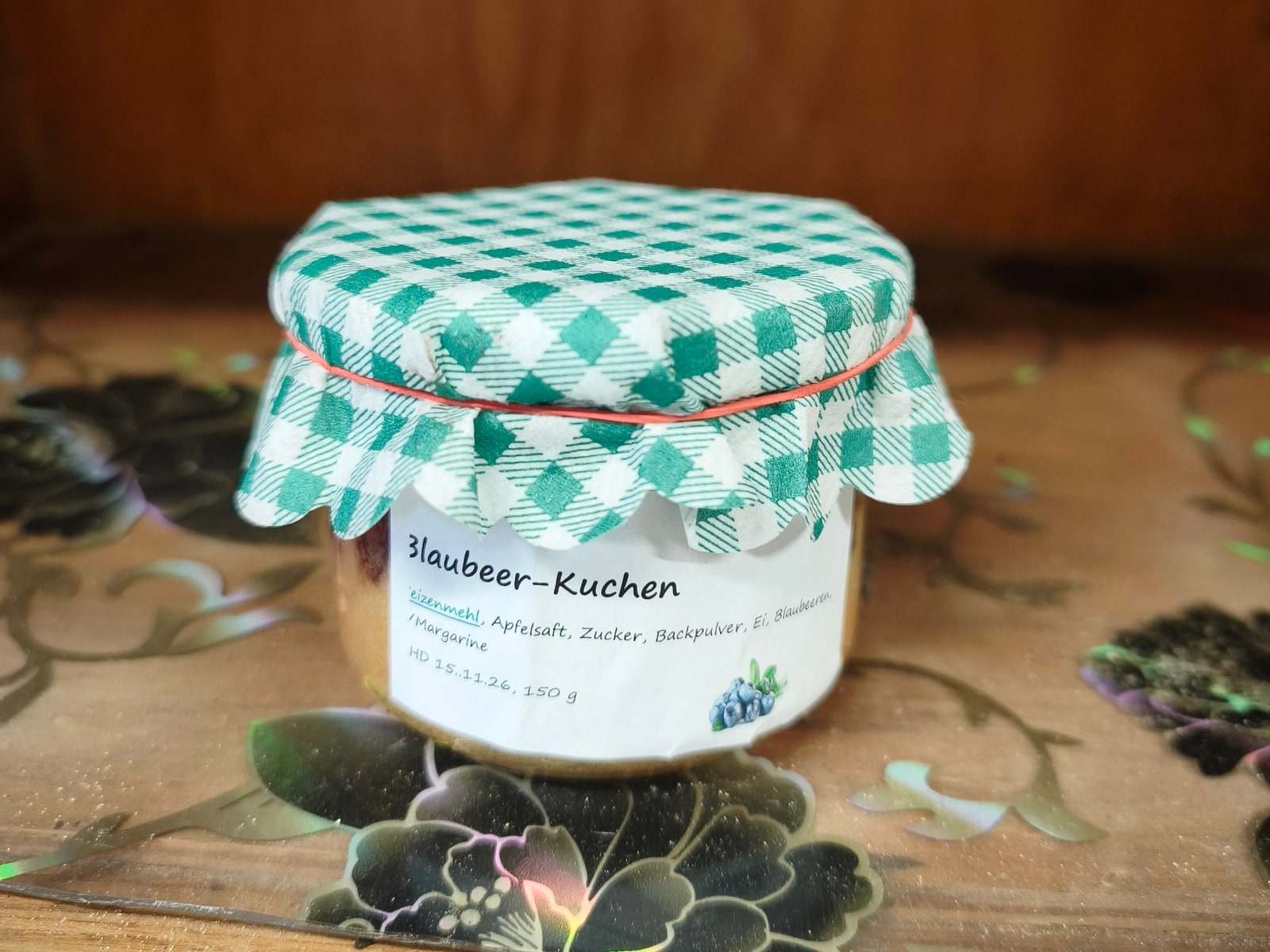 Ein Einmachglas mit Blaubeer-Kuchen steht auf einem gemusterten Brett im Verkaufswagen, über den Deckel ist ein grün-weiß kariertes Stoffdeckchen gebunden, davor ein handbeschriftetes Etikett.