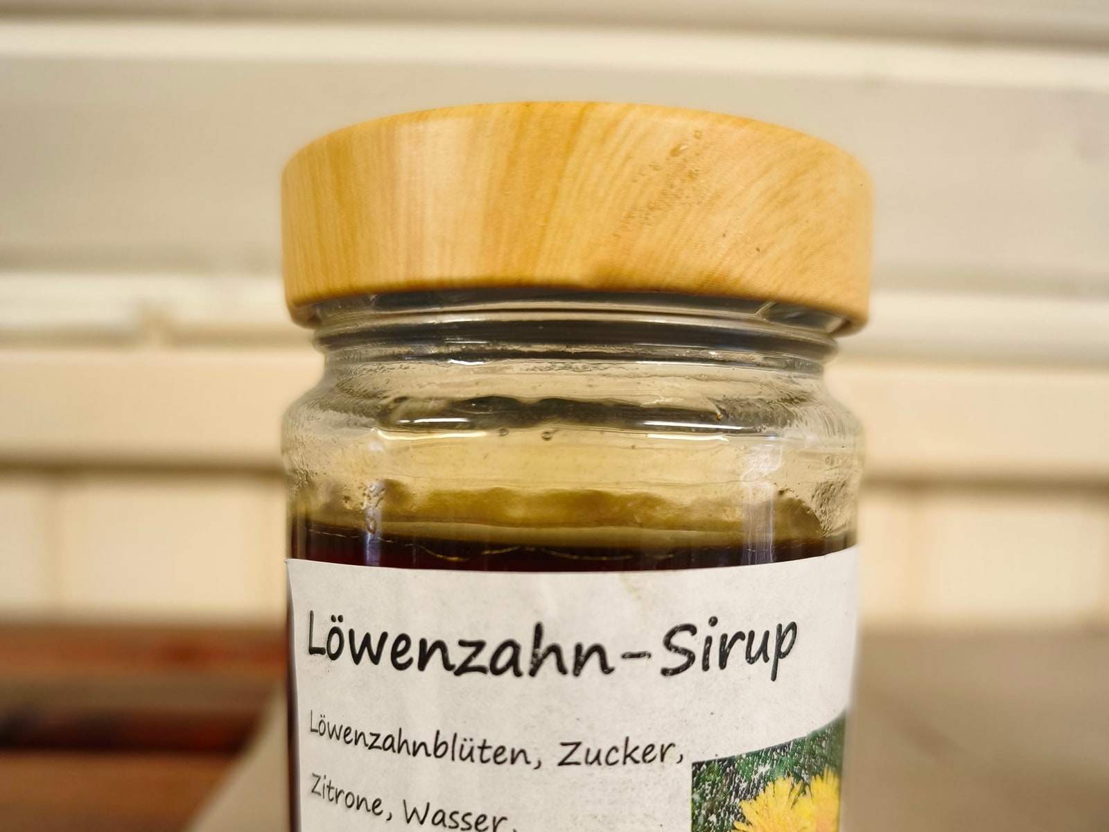 Ein Glas Löwenzahnsirup mit hellem Holzdeckel steht auf einem Brett im Verkaufswagen; auf dem handbeschrifteten Etikett stehen der Name und die Zutaten, daneben ein kleines Bild einer Löwenzahnblüte.
