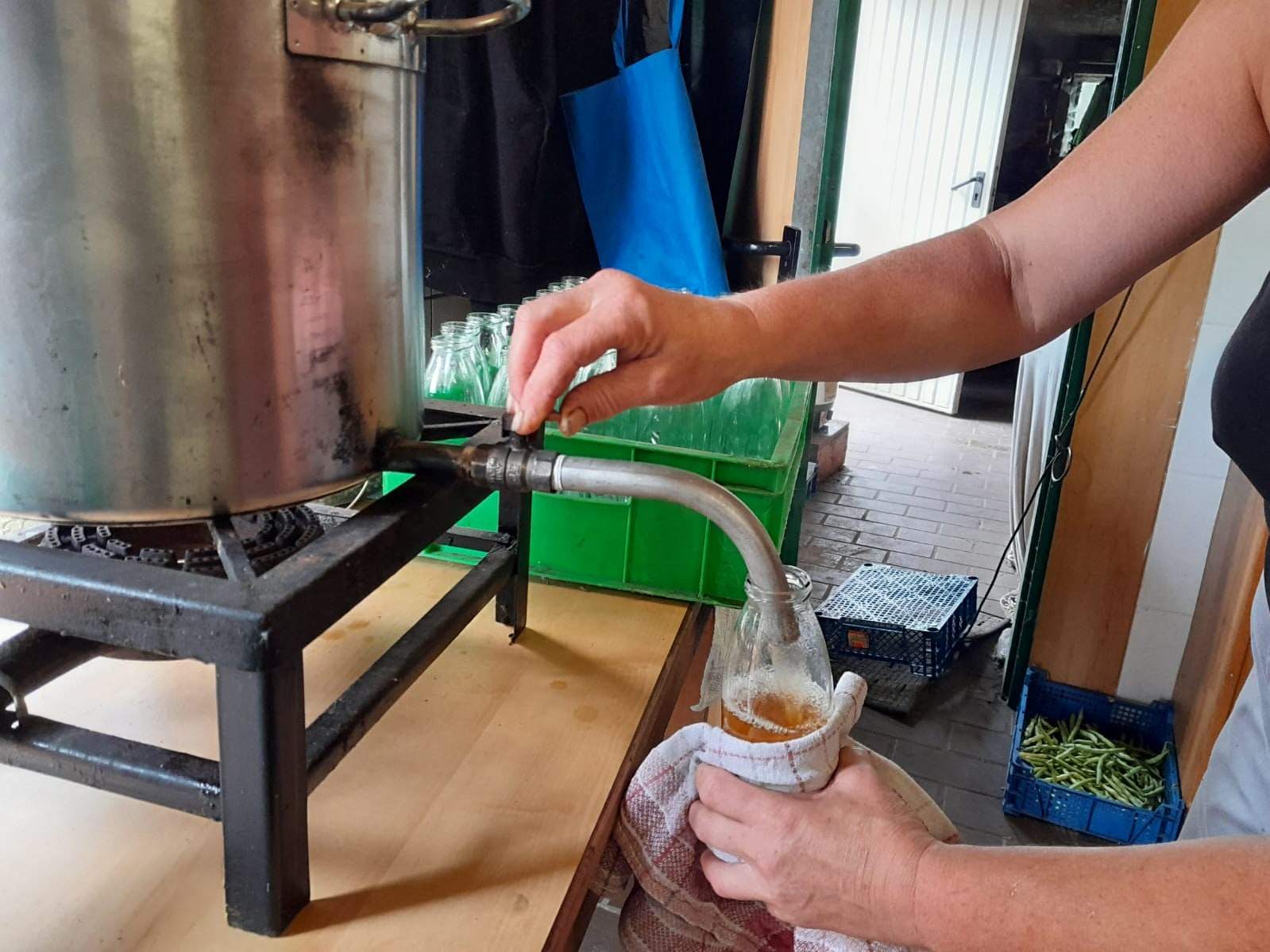 Zwei Hände am Hahn eines großen Topfes füllen heißen Saft in eine Flasche, die in einem Handtuch gehalten wird; dahinter eine Kiste leerer Flaschen.