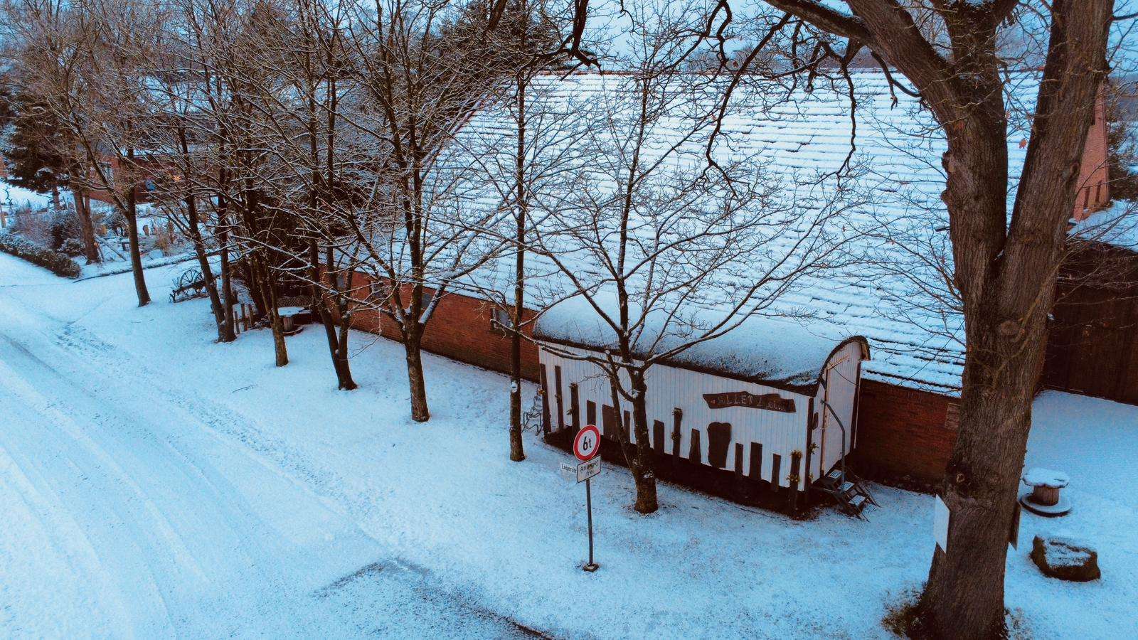 Blick von oben auf den weißen Verkaufswagen an der Klinkerscheune, ringsum Schnee, kahle Bäume und die Straße am Hof.